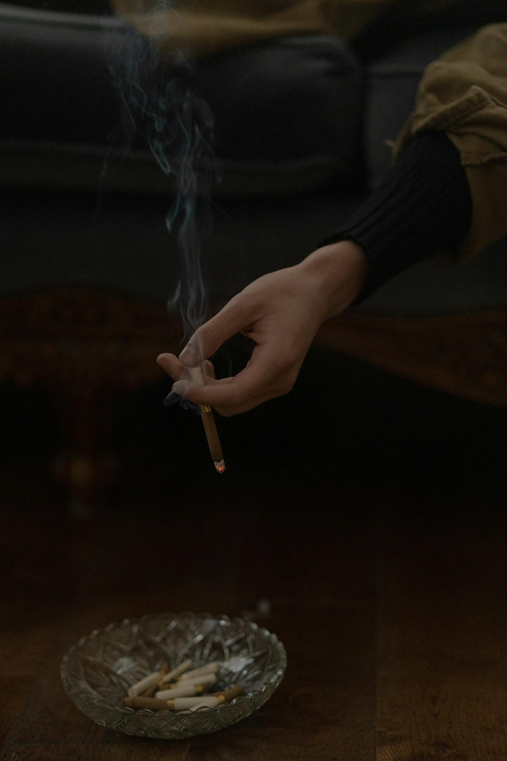 A hand holding a lit cigarette over an ashtray indoors, representing smoking habits.